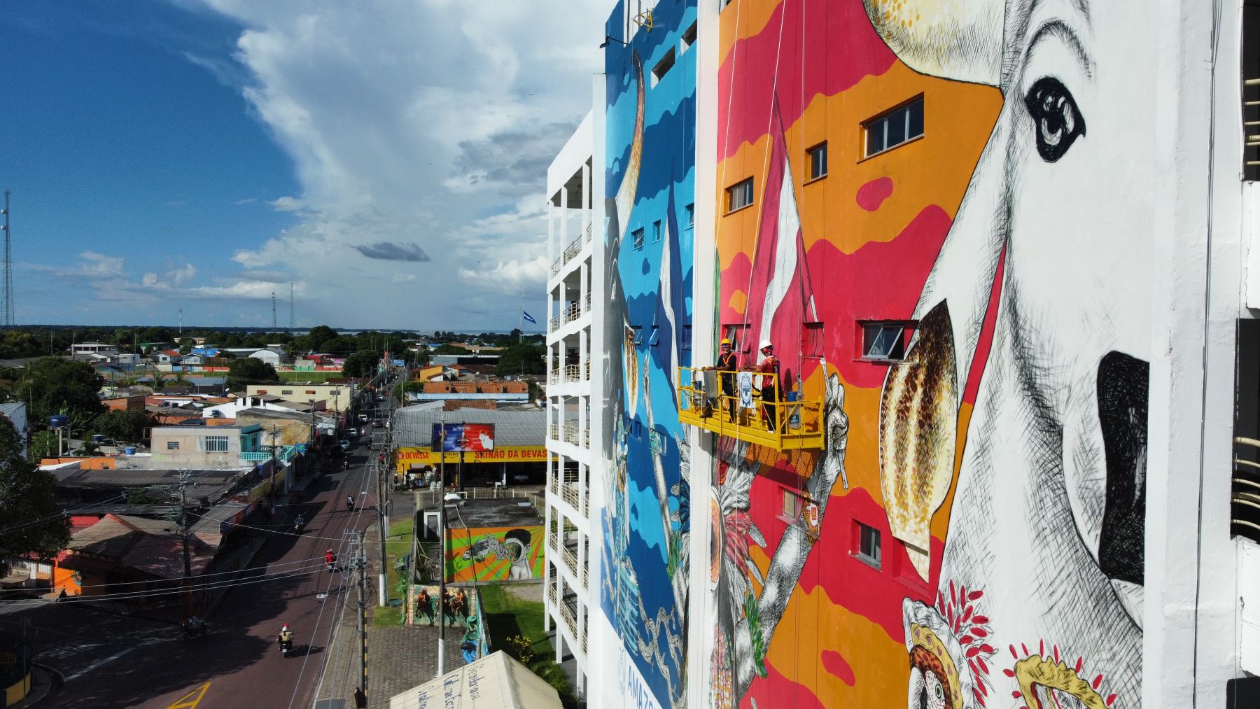Daqui a um mês, a fachada do Bumbódromo terá estampada a obra “Patrimônio em Festa”, de Pito SilvaUm canteiro de obras diferenciado com cores, artistas, tintas e grafite. A hora é de arregaçar as mangas e iniciar os trabalhos de pintura da fachada do Bumbódromo de Parintins (distante 369 quilômetros de Manaus). Nesta quinta-feira (02/05), o time de artistas liderado por Pito Silva, assume a missão de tornar visível a marca oficial do 57º Festival de Parintins que será estampada no complexo cultural.
Após receber as instruções de segurança e de estrutura dos equipamentos de suspensão, a equipe formada por mais de 20 artistas parintinenses, se dedica a dar vida à obra “Patrimônio em Festa”, de Pito Silva, contemplado no edital Mural Bumbódromo 2024, do Governo do Estado, por meio da Secretaria de Cultura e Economia Criativa.
Conforme o artista, todo o trabalho vem sendo cumprido dentro do planejado para que a entrega aconteça no período de um mês. “No primeiro momento, iniciaremos com a pintura civil, preparação da superfície da fachada para receber a nova arte. Na sequência, sobe a equipe de artistas visuais que estará fazendo a ampliação do projeto da arte para compor o mural deste ano”, detalha Pito.
A área total que será contemplada compreende 1.091 metros quadrados, incluindo fachada principal e as rampas dos bois Caprichoso e Garantido. Está prevista a utilização de 550 litros de tintas na composição, que mistura diferentes técnicas.
“A partir de ontem iniciamos todo esse processo de estudo e orientação de treinamento para realizar o trabalho com segurança. E quinta a equipe estará subindo no mural para a pintura da nova fachada do mural 2024”, reforça.
Conteúdo artístico
A arte “Patrimônio em Festa” tem a curadoria de Diego Omar, e traz a representatividade da cultura local, de modo especial ao folguedo popular do Boi, que ganhou novos sentidos na Amazônia, ao estabelecer cores e símbolos aos bumbás. O local onde será estampada a obra, não poderia ser mais emblemático que o Bumbódromo, templo do torcedor, representado por um curumim, símbolo da criança amazônica, convidando os visitantes para os dias de festa, 28, 29 e 30 de junho.
A nova arte irá ocupar o lugar da obra “Vitória da Cultura Popular”, da dupla Curumiz, por Alziney Pereira e Kemerson Farias. Nos últimos dois anos, Pito Silva é um dos artistas parintinenses do segmento de artes visuais atuante no circuito bovino. Ele assinou o mural “Brasil, Futuro Ancestral”, no Bumbódromo, na entrada da avenida Paraíba, por onde os bumbás se concentram para entrar na arena.
